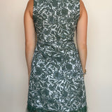 A woman viewed from the back wearing a sleeveless knee-length dress with an allover floral pattern in dark green and white, finished with fringe trim at the hem, against a plain light beige wall.