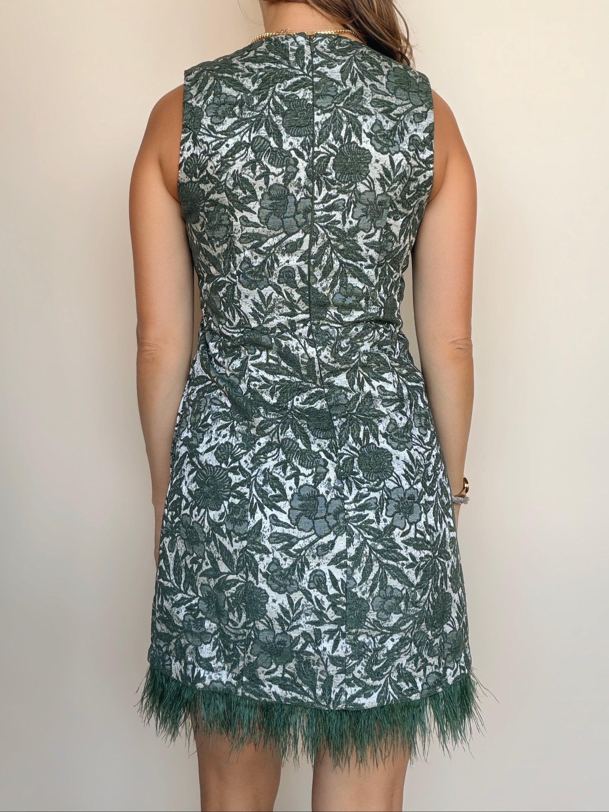 A woman viewed from the back wearing a sleeveless knee-length dress with an allover floral pattern in dark green and white, finished with fringe trim at the hem, against a plain light beige wall.