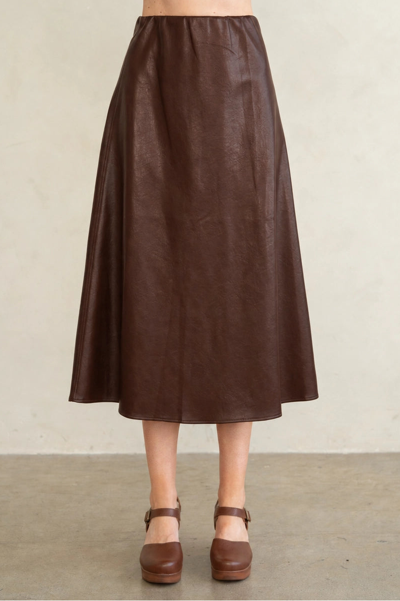 A model wearing a dark brown faux-leather midi skirt, paired with a matching dark brown platform Mary Jane shoe and a dark purple cropped top, standing against a neutral beige wall.