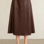 A model wearing a dark brown faux-leather midi skirt, paired with a matching dark brown platform Mary Jane shoe and a dark purple cropped top, standing against a neutral beige wall.