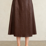 A model wearing a dark brown faux-leather midi skirt, paired with a matching dark brown platform Mary Jane shoe and a dark purple cropped top, standing against a neutral beige wall.
